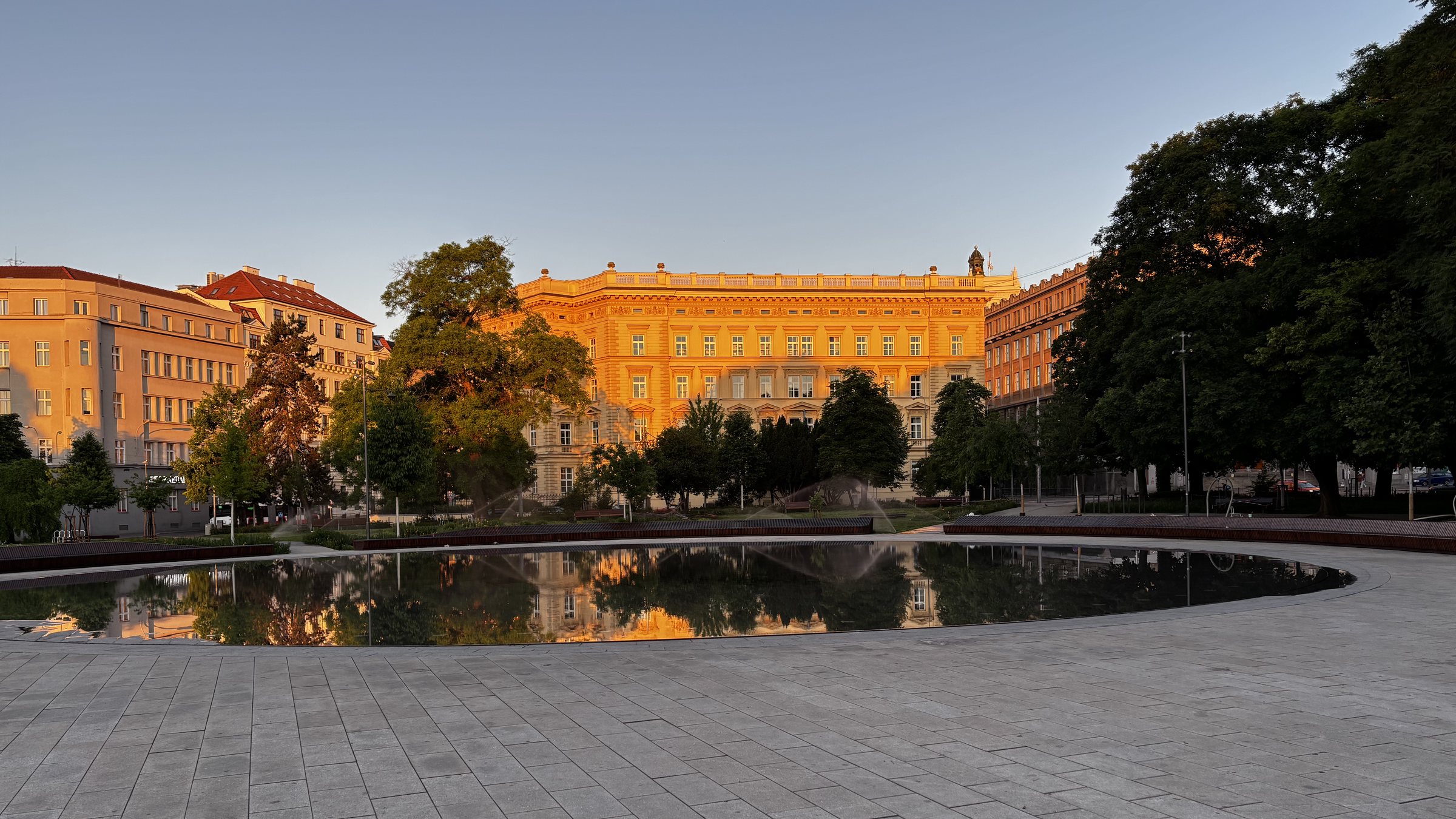 A dawn-lit Masarykova univerzita reflected off the Brněnské moře. Park sprinklers, flowers, the university building reflected on the pond, some lighting, an electric bus, trees and park benches, bookshelves in windows.