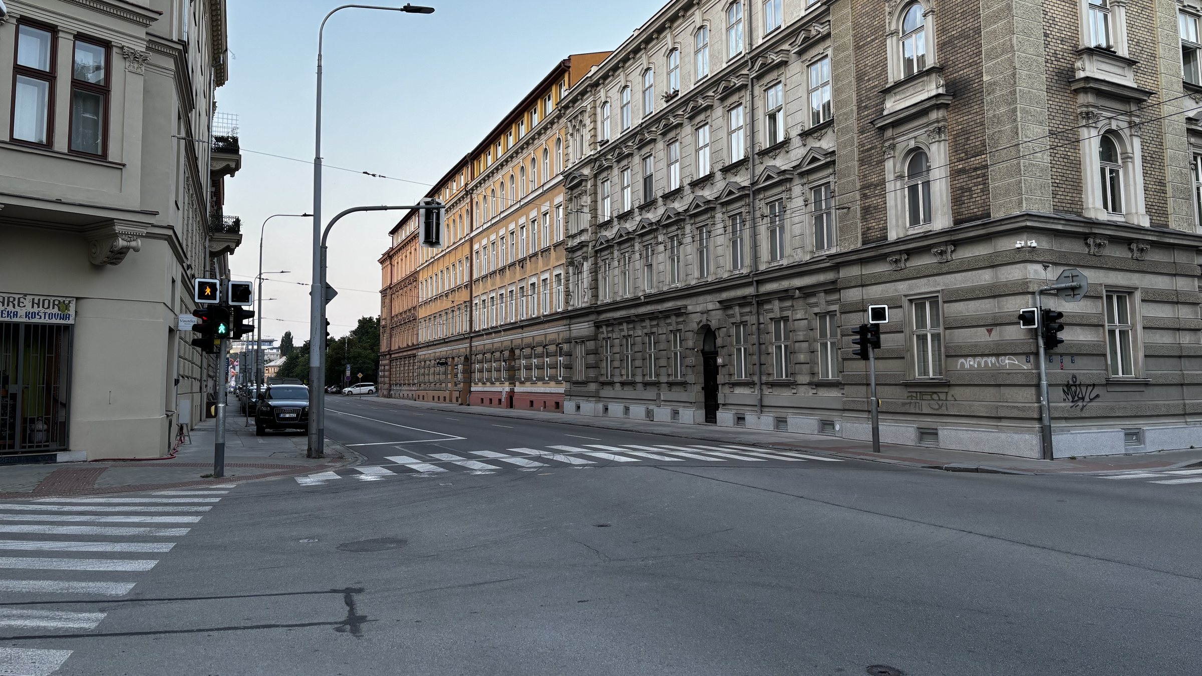 Intersection I. Varied facades in the predawn light, a green man, an orange man, Vinotékn, plants on balconies, catenary wires and an Audi, traffic lights and security cameras, window boxes, red power cable, grass and a gas bottle in a window.