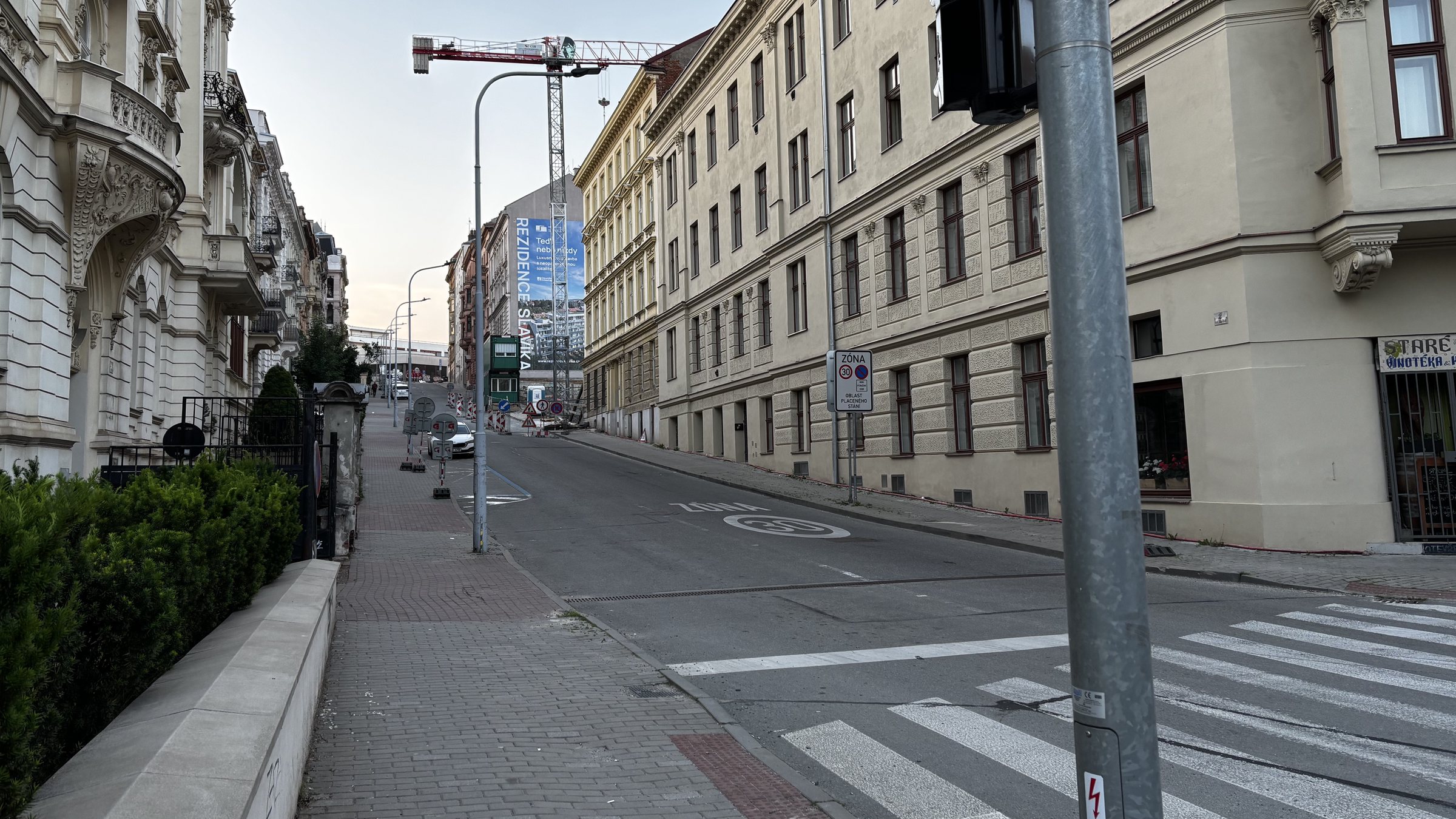 Intersection II. Front gardens beneath ornate balconies, person walking with a bag, temporary signs, graphiti, site huts, and a keep left and road narrows sign. Flowers in window, Vinotéka, a gate catch and yesterday's specials. A big red power cable runs through grass under a bent sign. Rezidence Slavika behind a jack up crane.