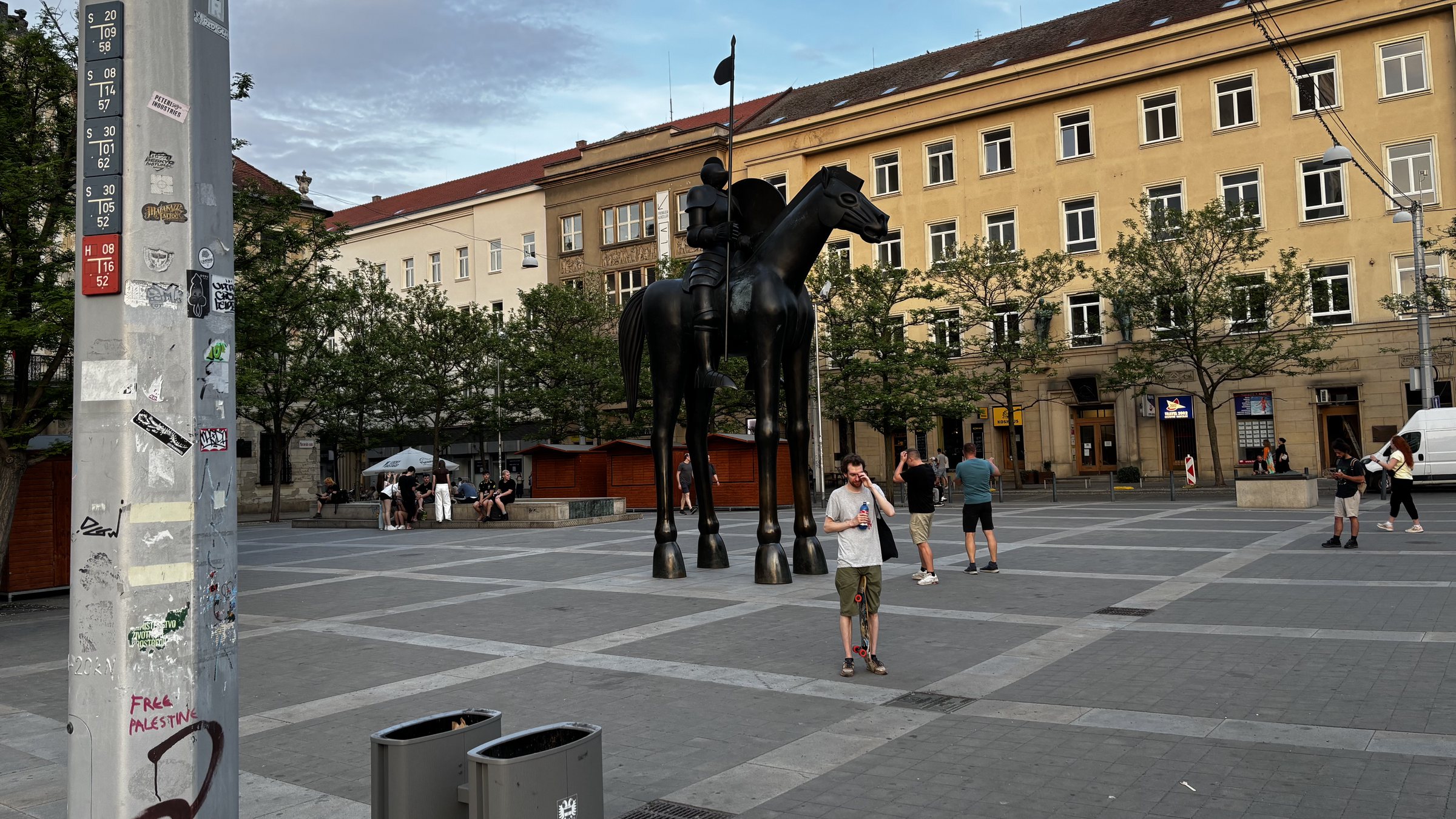 Socha markraběte Jošta Lucemburského. Catenary support pole with tram specific markers on it, band and club stickers, and some Free Palestine graphiti. Young people enjoying the evening and a guy rubbing his eyes while holding a skateboard between his legs. A building with a carved facade, overhead suspended lighting, a ladder in a door and a red safety bollard, dude with phone a brass plaque and a discarded spoon.