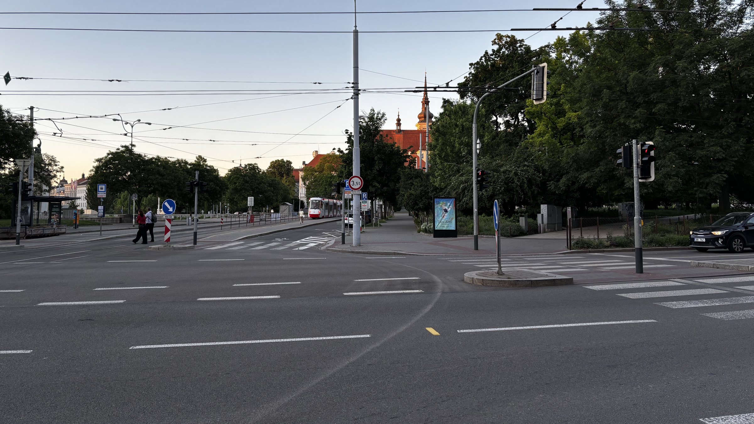 Lidická and Koliště. Traffic light, a skip bin, a statute with a fist held high, tram stop, tram tracks, keep right, a tram, Den dětí, mesh fence, graphiti, a church and its steeple, electrical boxes, grass and flowers, reserve parking and catenary wires.