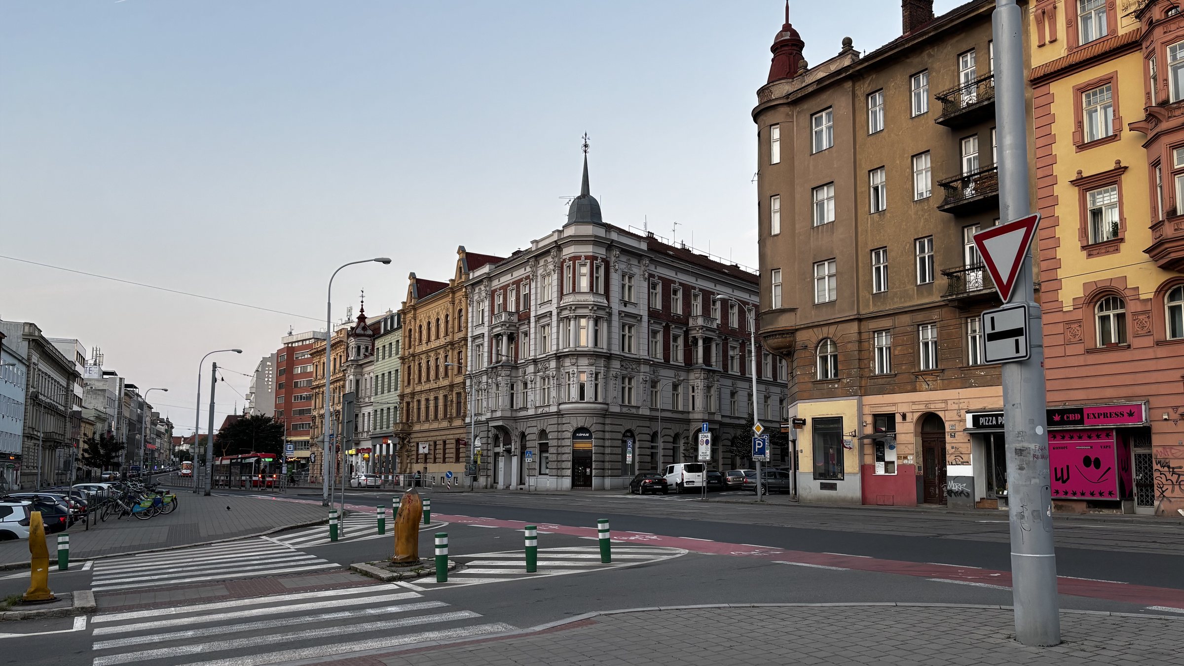 Varied buildings, varied history at dawn. Give way (yield) sign, perspective, trams on the tram lines, bollards, vape shop, pizza shop, open window with a plant, balcony with a shipping palette on it, crumbling render, a statue of a man in the window, a brass dome, antennas, brick render, lions, faces, laurells, Nápoje, colums, gables, keep right, man at tram stop, hotel, e-bikes, neon, grass.